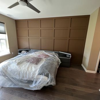Modern bedroom with brown paneled accent wall, bed with plastic covering, hardwood floors, and ceiling fan
